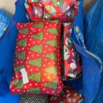A blue shopping bag is full of presents wrapped in red Christmas themed wrapping paper. They are all ready to be delivered to underserved youth.
