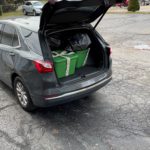 A black car with an open trunk. In the trunk there are presents that are wrapped in green paper and ready to be delivered to underserved youth in the area.